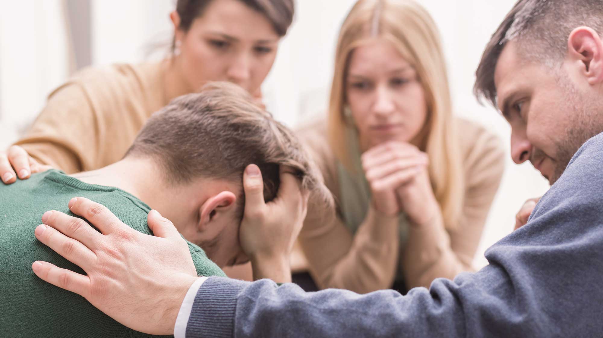 Close-up of a devastated young man holding his head in his hands and friends supporting him during group therapy